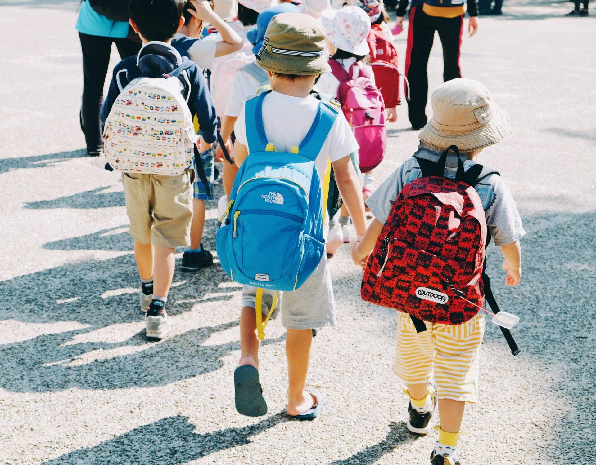 Photograph of young children walking outdoors, each wearing colorful backpacks and sun hats. Bright sunlight casts shadows on pavement.