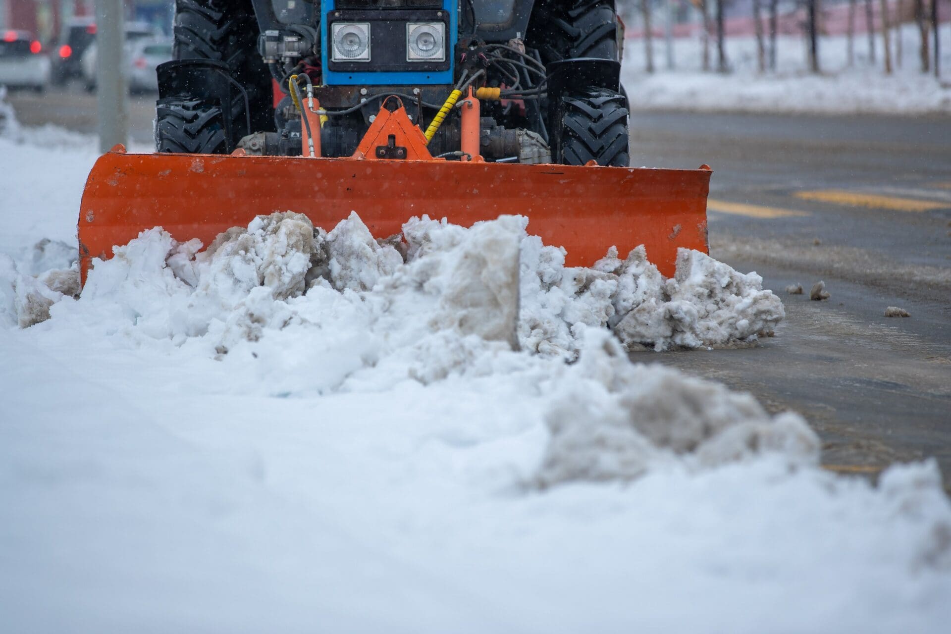 Photograph of a snowplow clearing snow from a street, showing an orange plow blade pushing accumulated snow to the side.