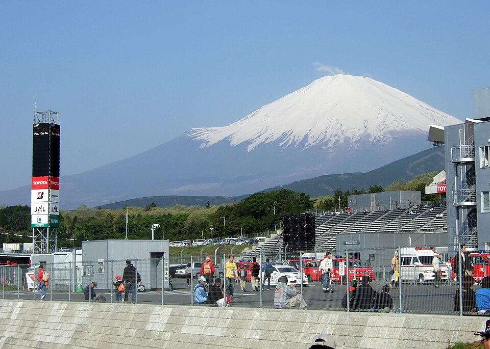 Photograph of Mount Fuji behind a motorsport racetrack, showing pit buildings, grandstands, fencing, officials, spectators, and support vehicles. Snow-covered peak dominates background, while a tall timing tower and red-and-white track signage mark racing venue setting.