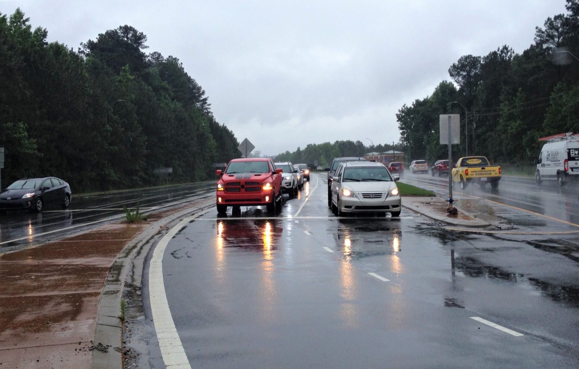 Photograph depicts rainy roadway traffic, with cars traveling along wet lanes beside dense forest. Headlights and taillights reflect across pavement.