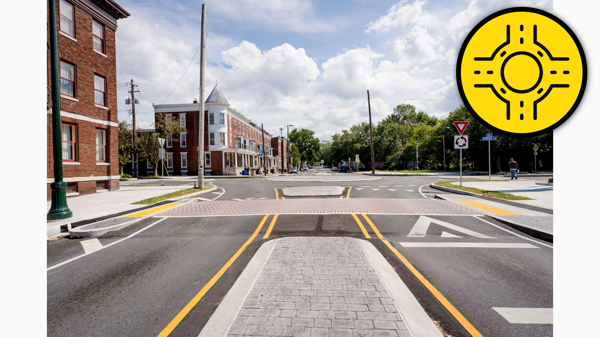 Photograph showing a city street roundabout intersection with brick buildings, sidewalks, trees, and a cobblestone traffic-calming median.