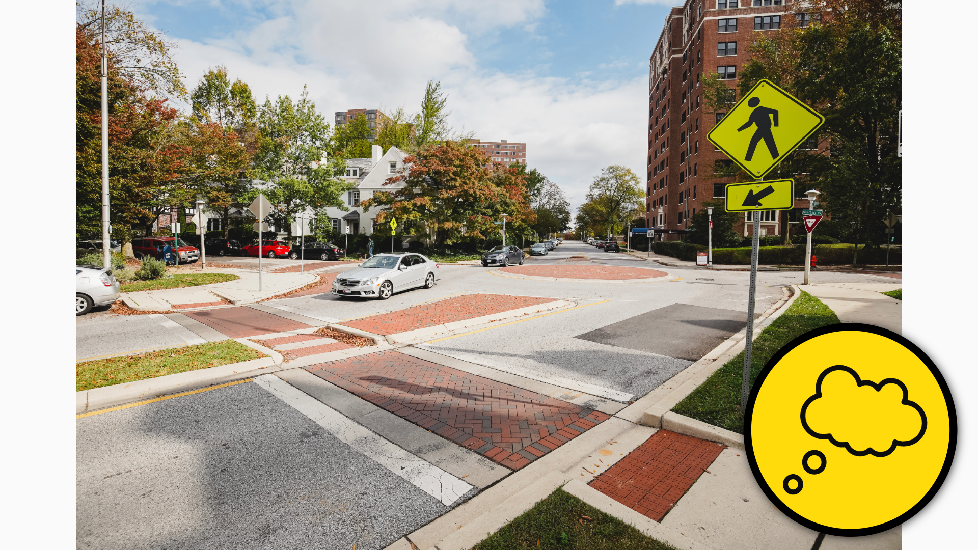 Photograph showing a roundabout intersection with brick-patterned pedestrian crossings. There are also yellow pedestrian-warning and directional signs, yield sign, cars, and curb ramps.