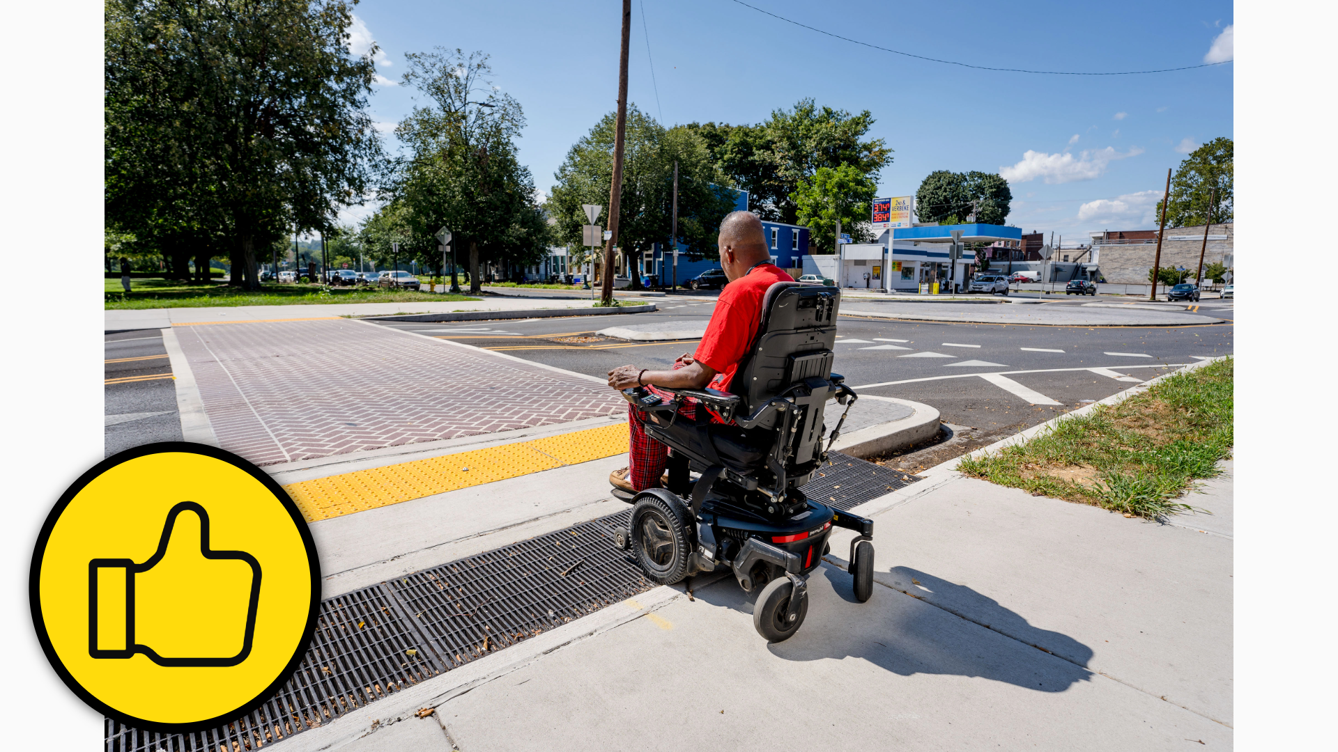 Photograph shows a person using a motorized wheelchair approaching a curb ramp at a roundabout intersection. There are yellow tactile paving, drainage grates, marked crosswalk, traffic signals and trees.