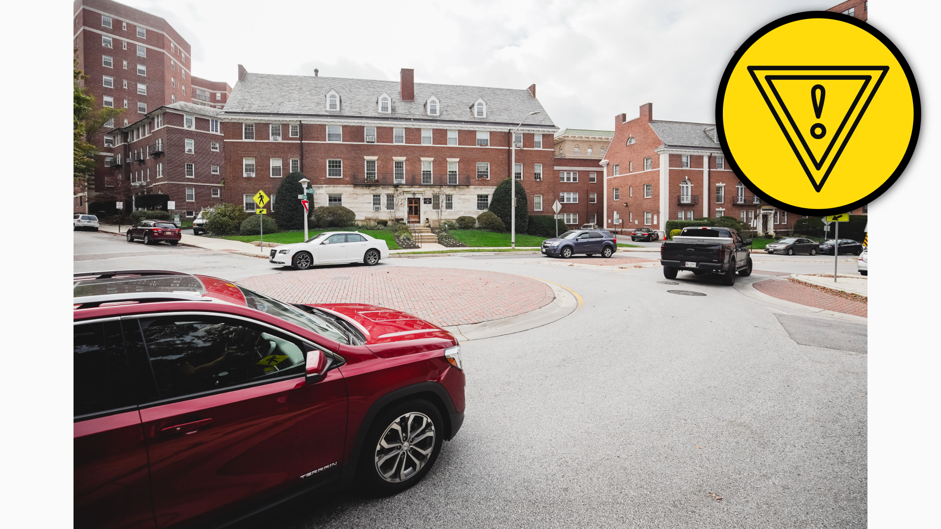 Photograph showing a landscaped roundabout, red-brick institutional buildings, high-rise apartments, parked cars, and a red SUV in foreground. Yellow pedestrian-crossing signs, crosswalk markings, and overcast skies.