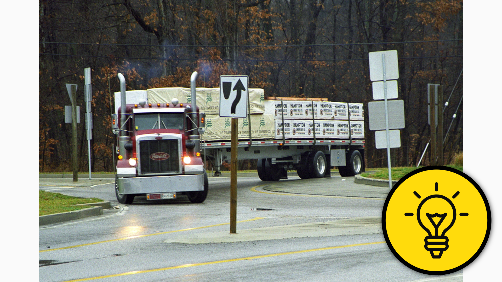 Photograph a semi-truck navigating a roundabout intersection beside a wooded area.