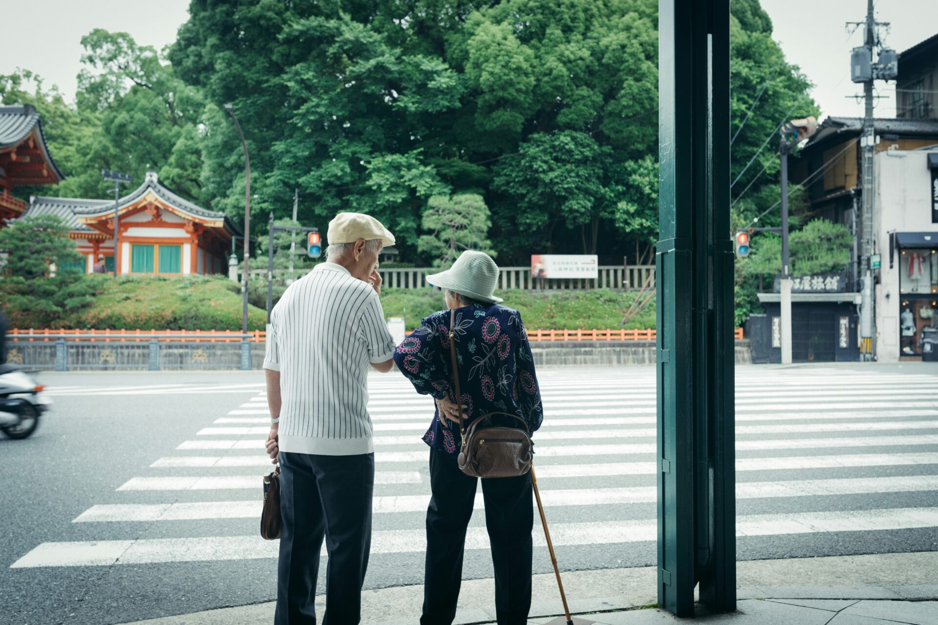 Photograph shows an older couple waiting at a zebra crossing beside a broad city street, with brightly painted Japanese architecture and lush trees in the background.