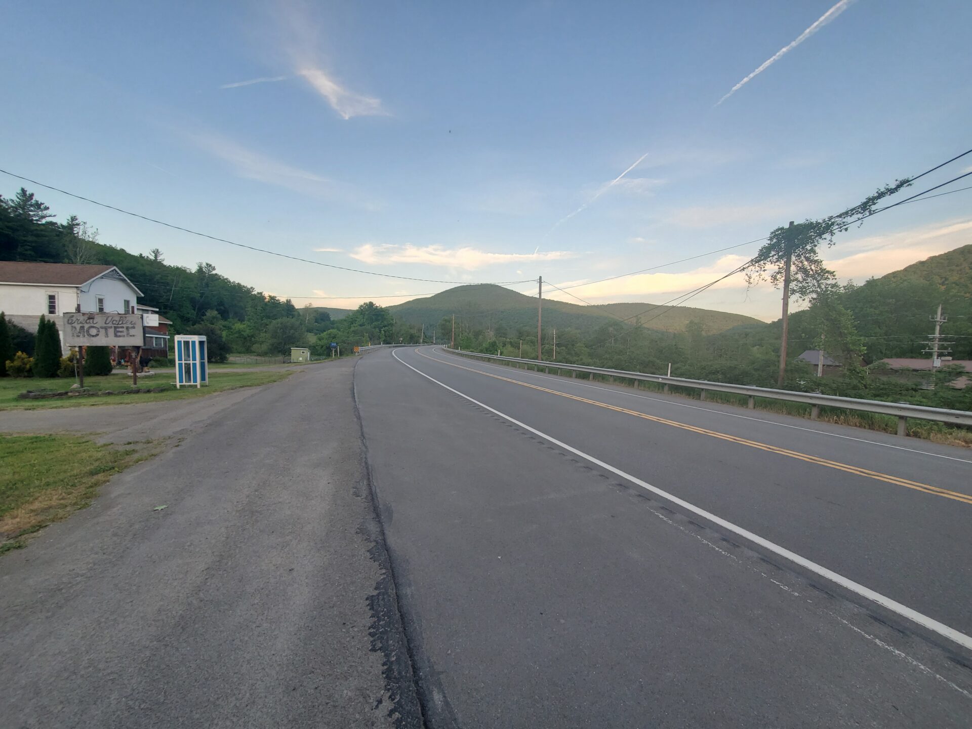 Photograph shows a rural two-lane highway curving through wooded hills, with double yellow centerlines, white edge markings, guardrail, utility lines, and roadside motel sign.