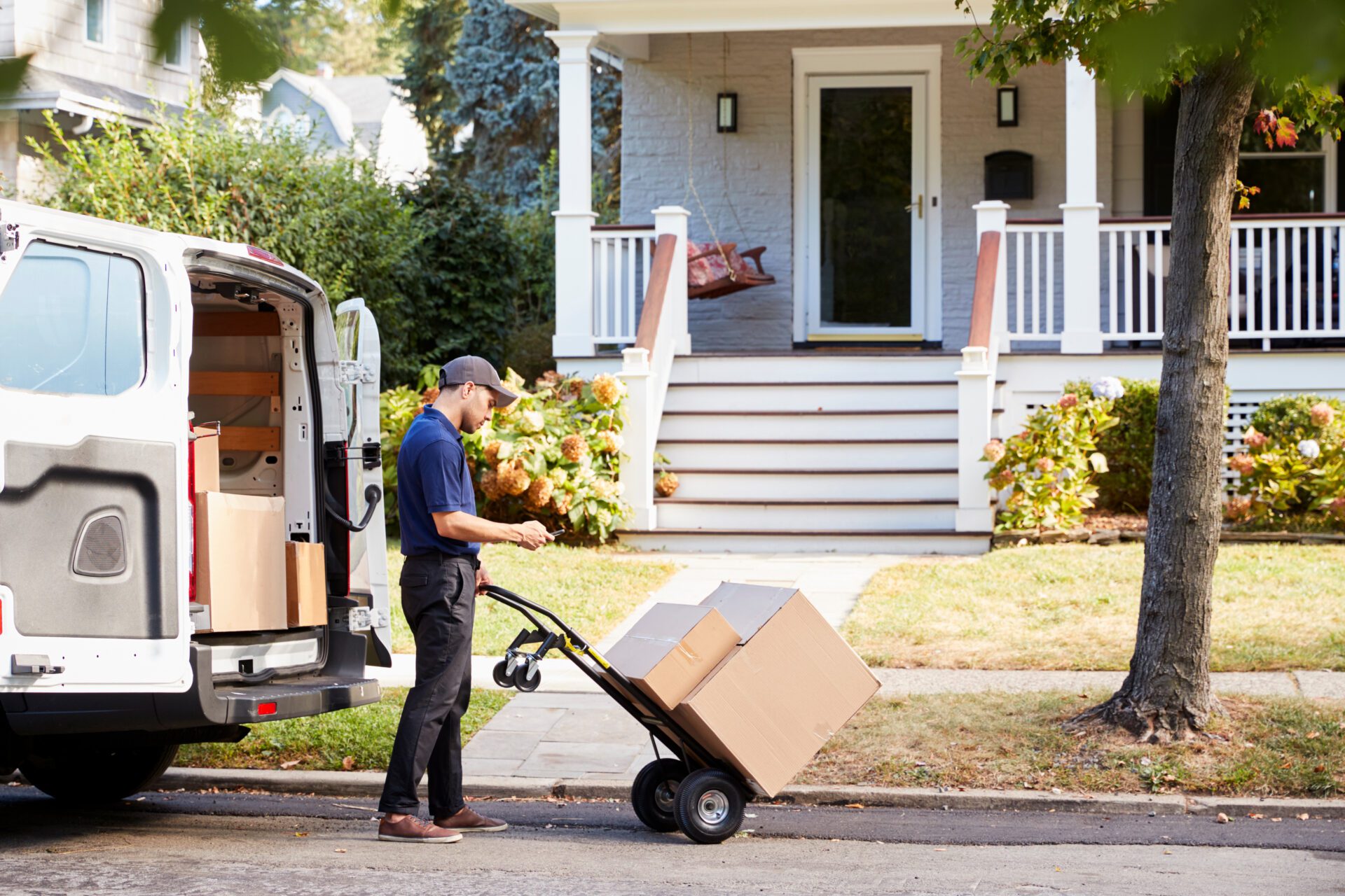 Delivery worker unloading packages from a van in front of a residential home. Several cardboard boxes are stacked on a hand truck on the sidewalk, with the delivery vehicle parked at the curb nearby.