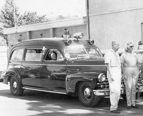 Black-and-white photograph shows a vintage black emergency vehicle with roof-mounted siren, lights, polished chrome, and a driver visible through side window. Two adults stand beside vehicle outside a building.