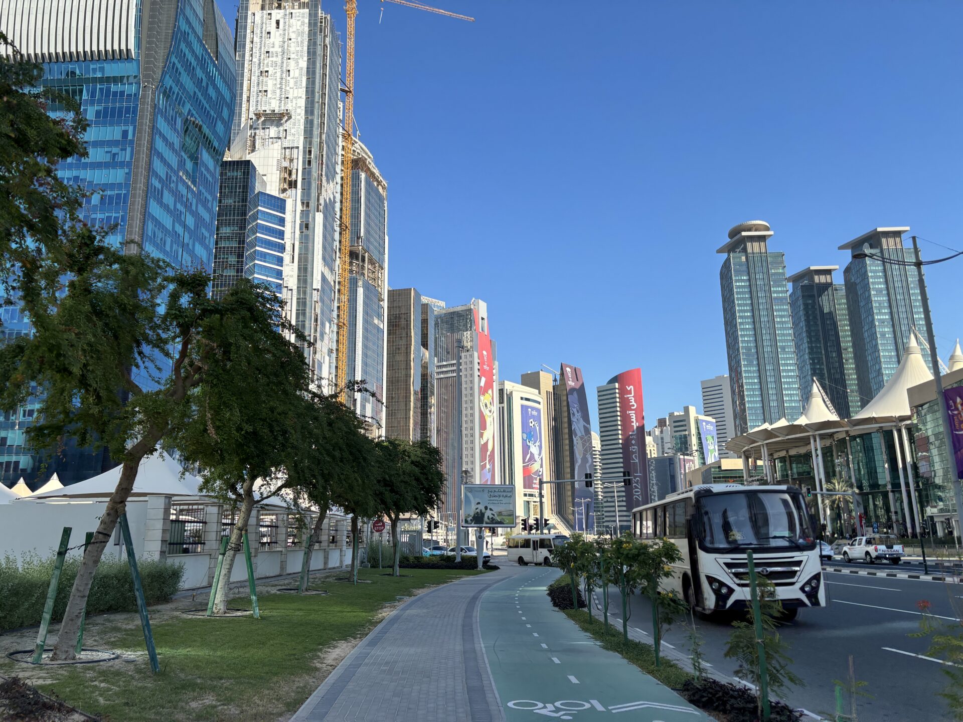 Photograph showing a modern urban boulevard lined with glass skyscrapers, construction cranes, landscaped trees, and a dedicated bicycle path. White city bus, multi-lane roadway, commercial towers, and clear blue sky highlight dense city development and active transportation infrastructure.