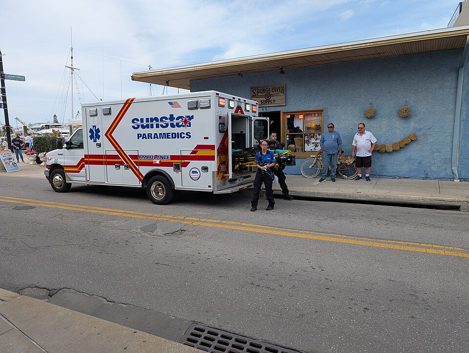 Photograph showing a Sunstar Paramedics ambulance parked outside a building with emergency personnel and bystanders nearby.