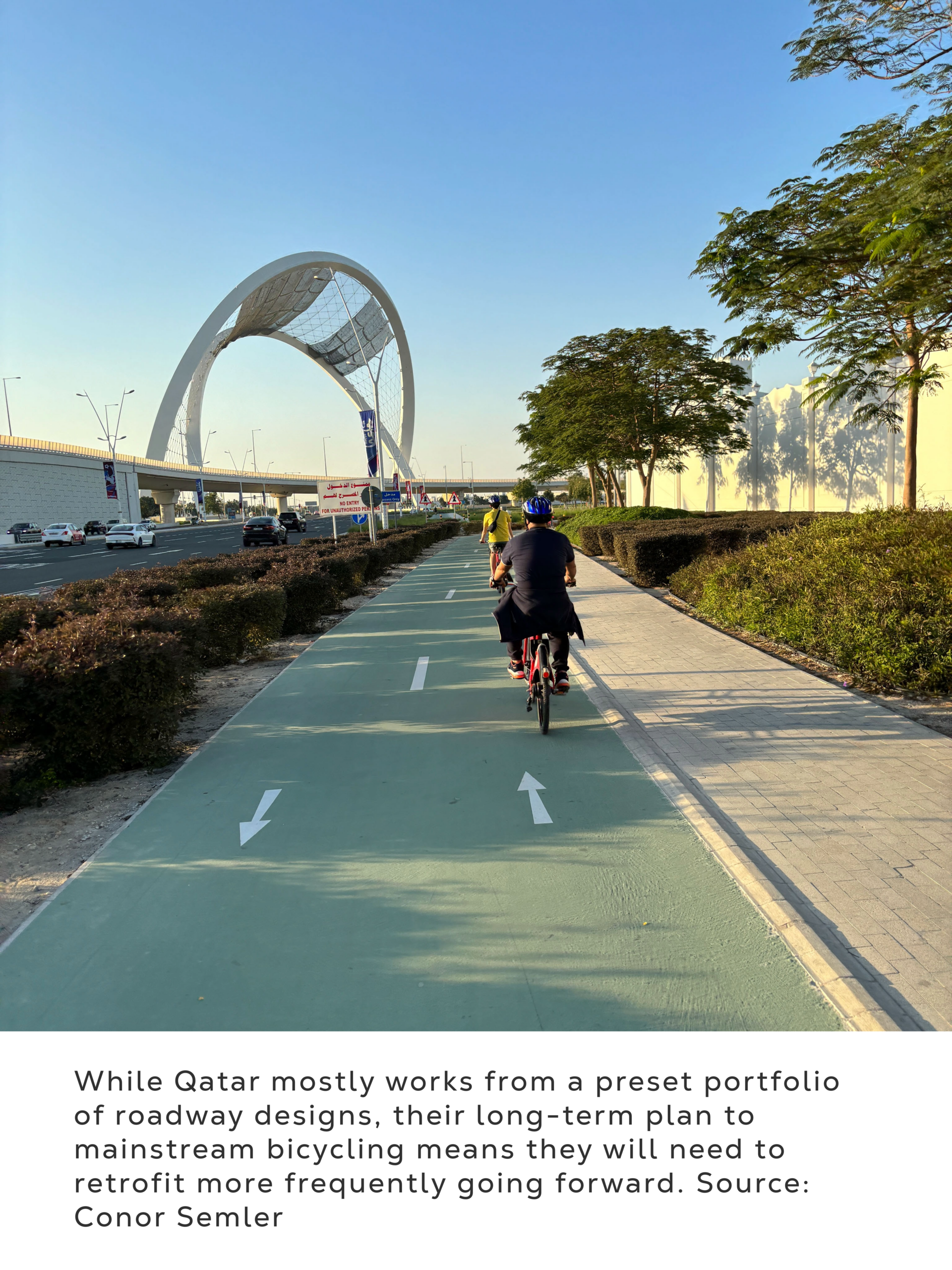 Photograph showing a cyclist riding along a green, marked bike path beside a roadway and landscaped hedges. White directional arrows, an arched pedestrian bridge, trees, streetlights, and distant buildings establish an urban transportation corridor.