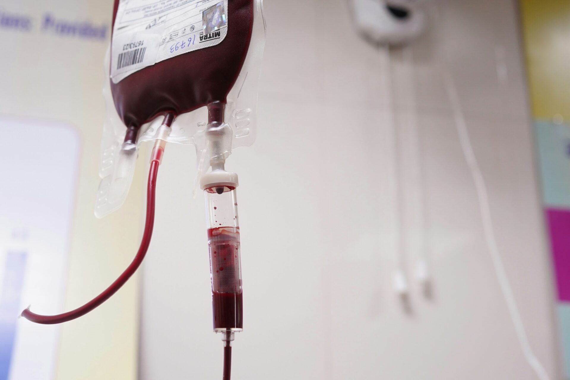 Photograph shows a dark red blood bag connected to clear tubing and a drip chamber in a hospital setting.