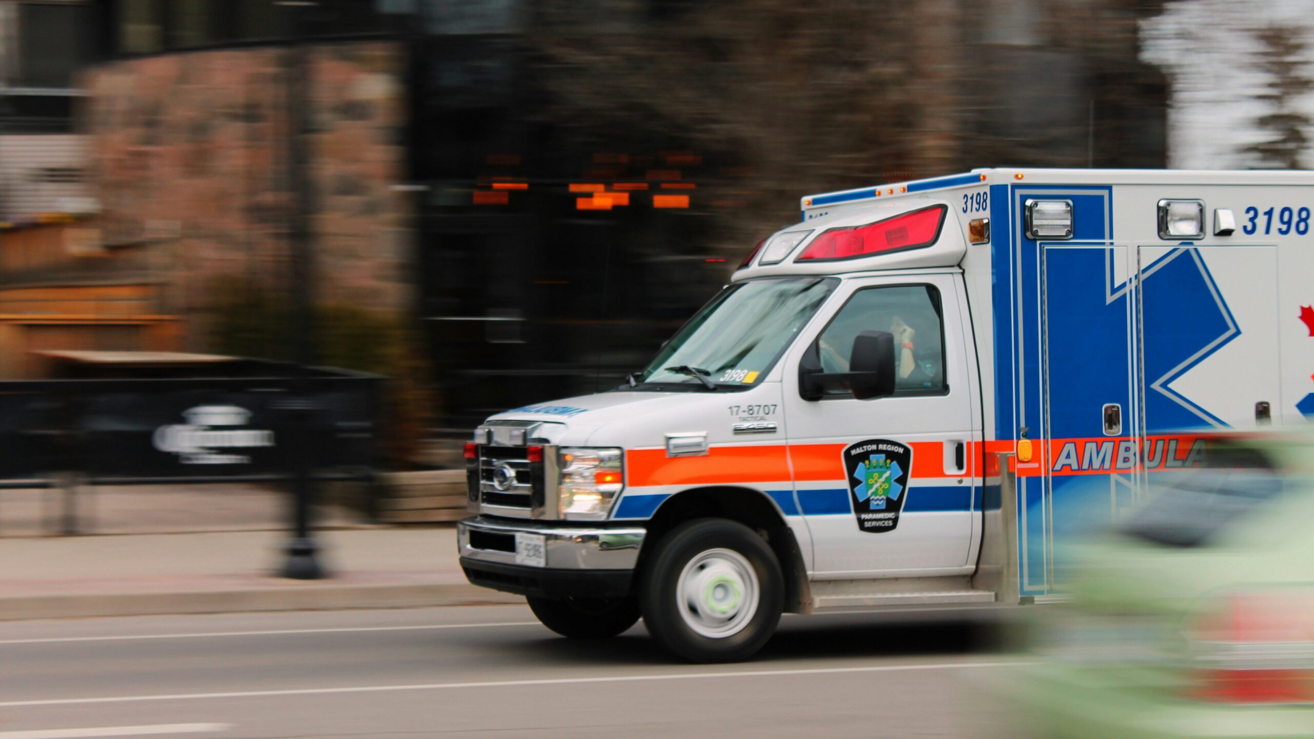Photograph shows a blue, white, and red ambulance speeding through an urban street during an emergency response.