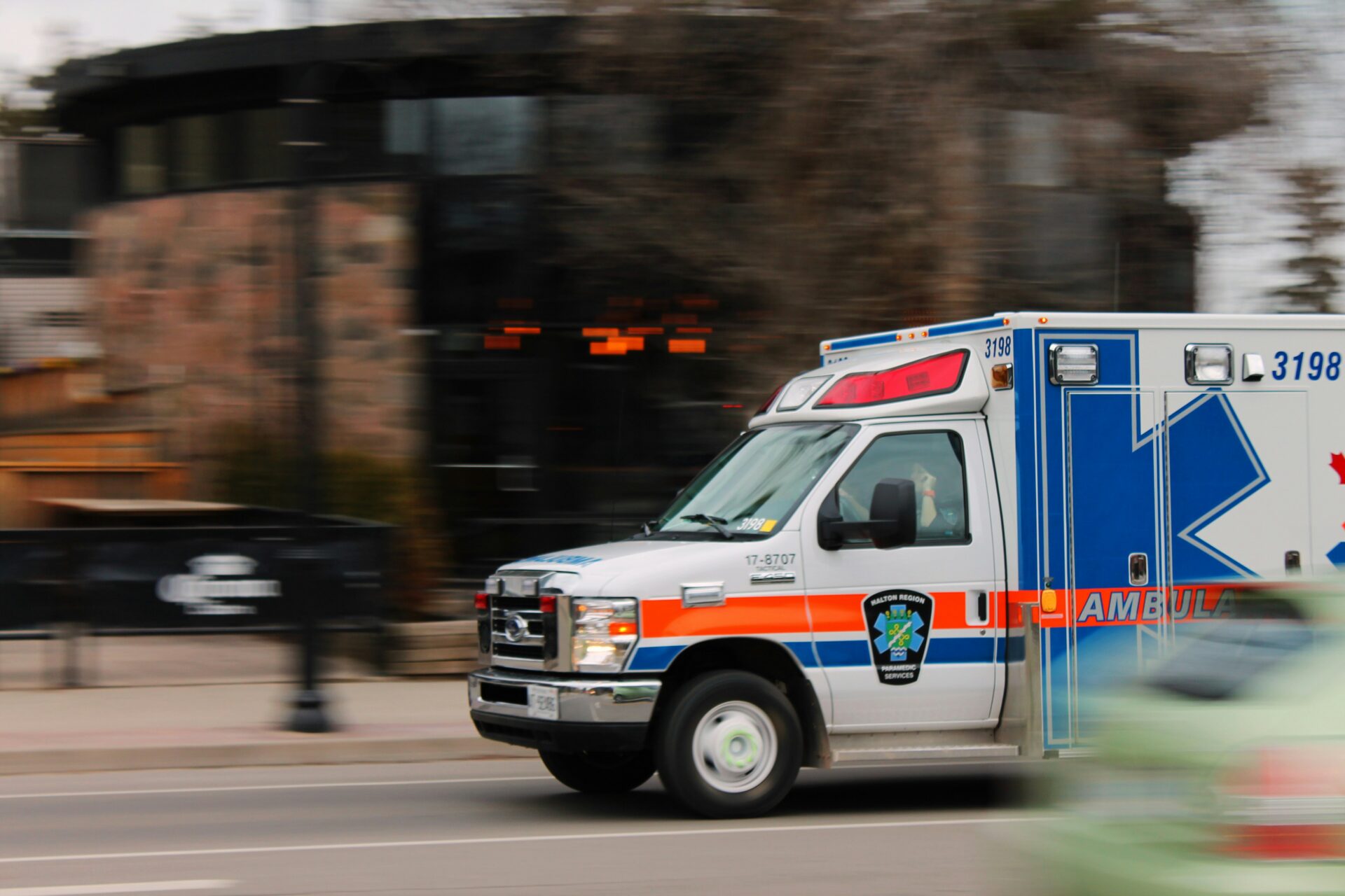 Photograph shows a blue, white, and red ambulance speeding through an urban street during an emergency response.